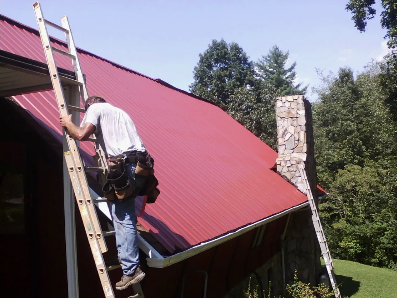 Roofer on a ladder beside a red metal roof and stone chimney — Soffit & Fascia Repair by Roofing Brothers in Binford